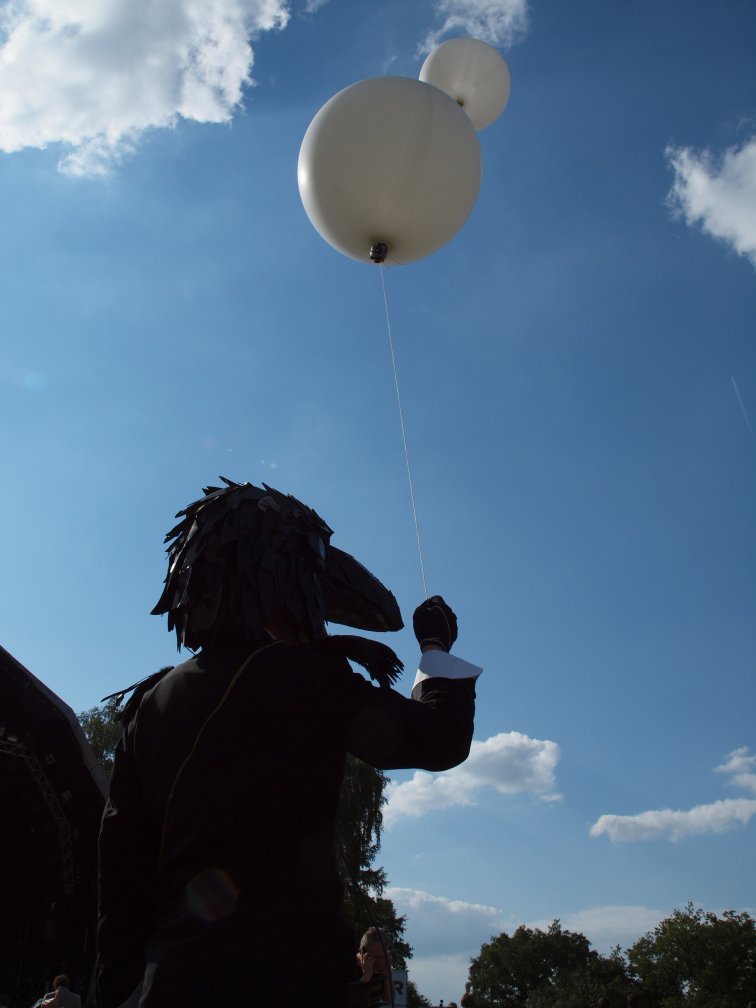 Man wearing a crow's head over his head holding two large white balloons